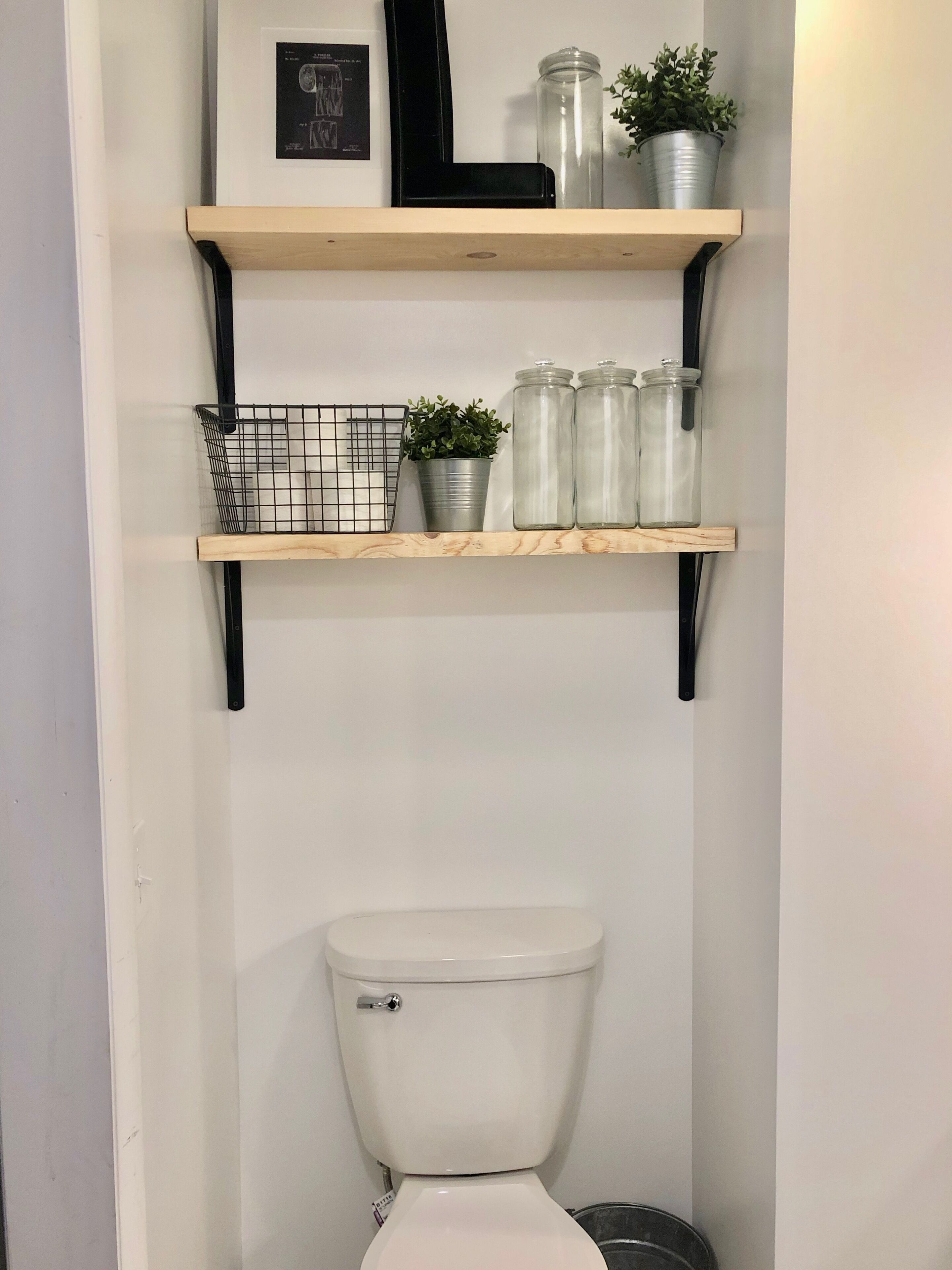 A bathroom with a toilet, two shelves with storage jars and a plant, and a metal basket.