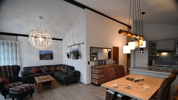 A modern living room with a sofa, coffee table, and dining table. There is a kitchen area with cabinets and a hanging light fixture. - Holiday home Kenter Tossens (Butjadingen)