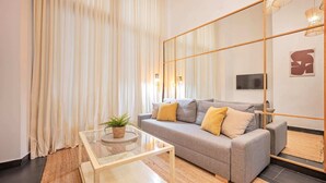 Una sala de estar moderna con un sofá gris, una mesa de centro de vidrio y un espejo grande que refleja la habitación.