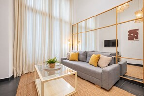 A modern living room with a grey sofa, yellow cushions, a glass coffee table, and a large mirror reflecting the room. - Sweett | Gothic Baixos (Barcelona)