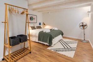 Une chambre à coucher avec un lit, un ventilateur sur pied, un étagère en bois avec des paniers et un tableau encadré accroché au mur.
