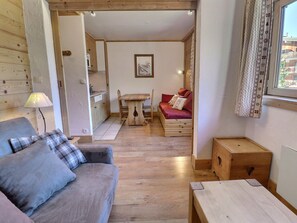 A cozy room with a sofa, a wooden table, a small kitchen area, and a window with curtains. - Charming Studio in Méribel - Ski-in/Ski-out, Pets Allowed (Méribel)