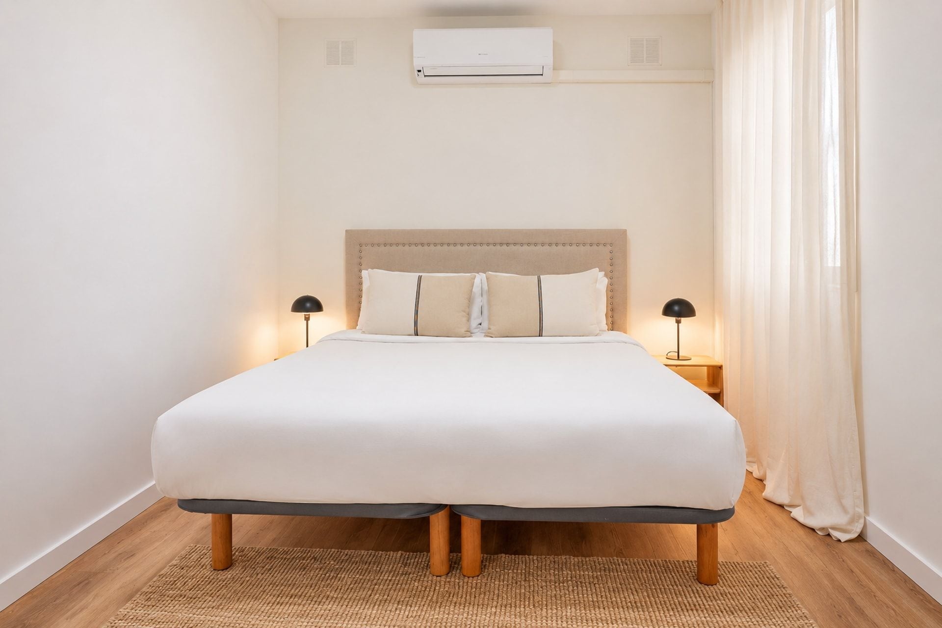 A bed with a headboard, two bedside lamps, a wall-mounted air conditioner, and a window with curtains.