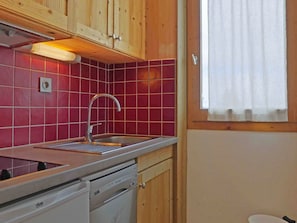 A compact kitchen with a stainless steel sink, wooden cabinets, and a window with a white curtain.