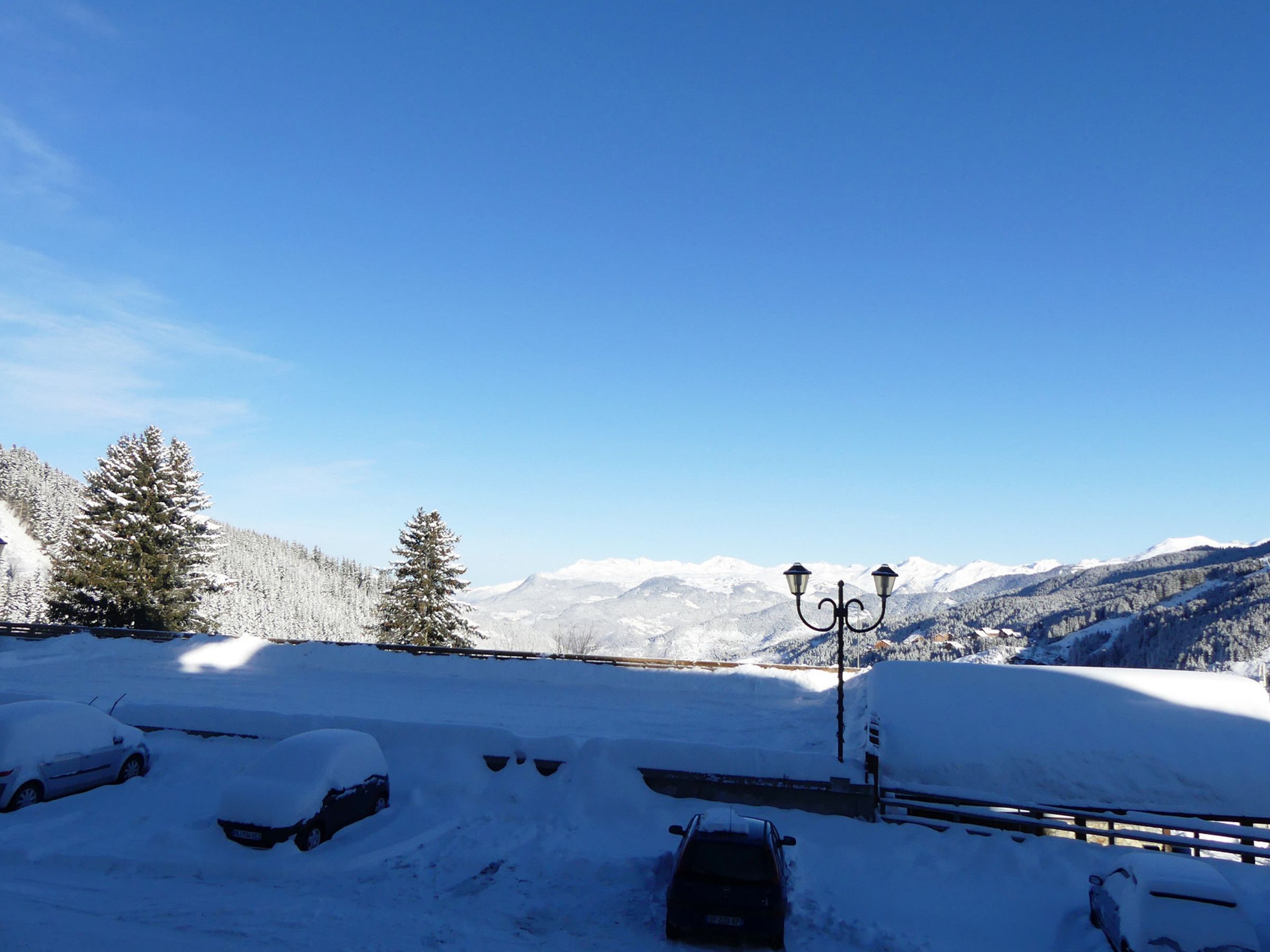 A snow-covered landscape with a bench and a lamppost, surrounded by snow-laden trees and mountains in the background.