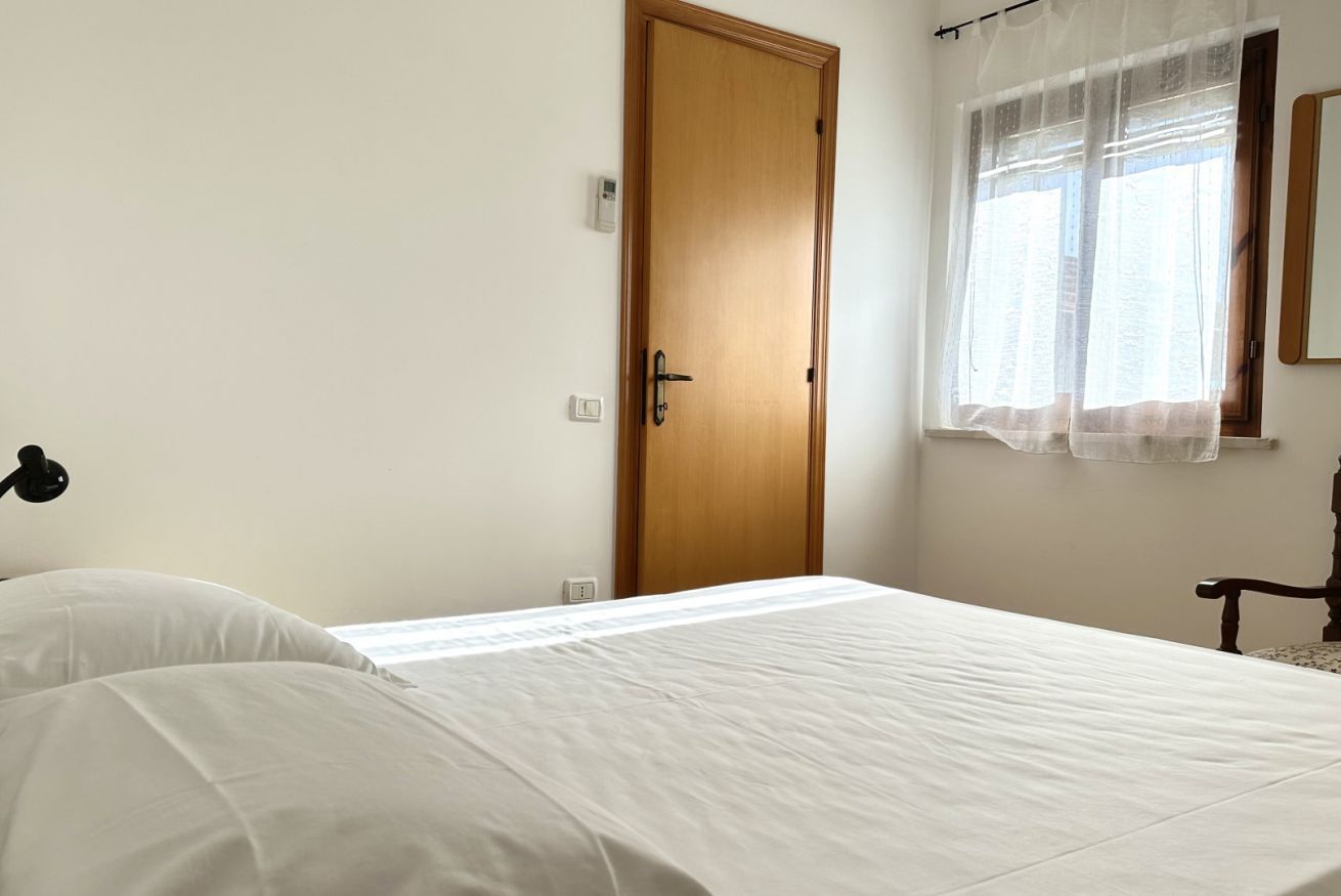 Un dormitorio con puerta de madera, una mesita de noche, una cama con ropa blanca, una ventana con cortinas traslúcidas y un espejo montado en la pared.