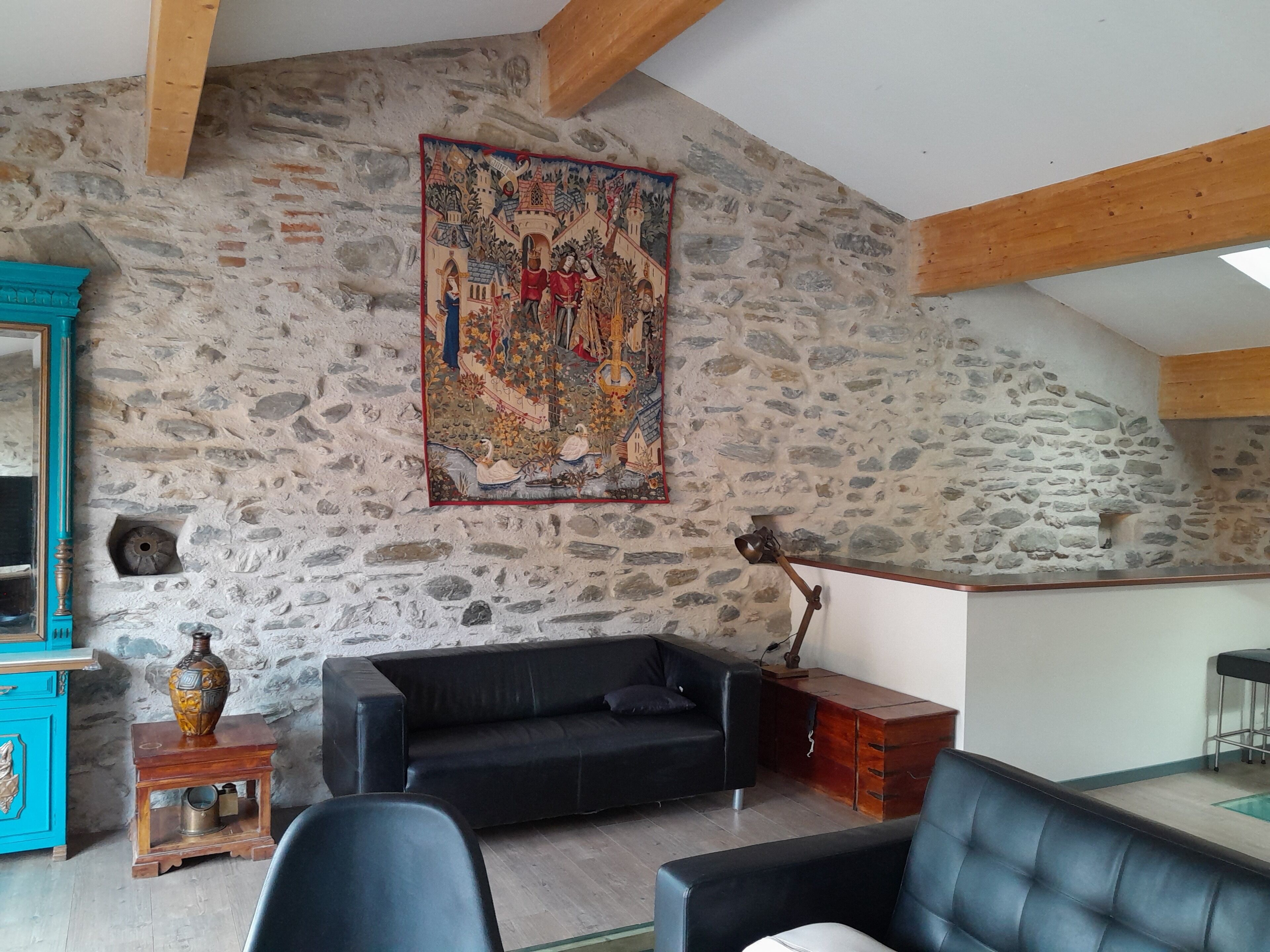 A room with a stone wall, a large tapestry, a black leather sofa, a wooden cabinet, and a teal cabinet.