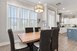 Un comedor con mesa de madera y sillas tejidas, una cocina con armarios blancos y una lámpara decorativa en el techo.