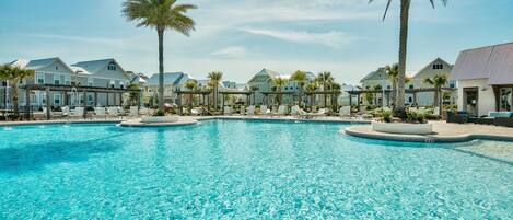 A large swimming pool with lounge chairs and palm trees, surrounded by resort-style buildings.