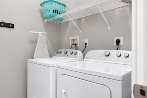 A laundry room with two front-loading washing machines, a white towel rack, a blue laundry basket, and a white shelf above the machines.