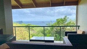 Un balcone con mobili in vimini, ringhiera in vetro e vista sull'oceano e sulla costa.