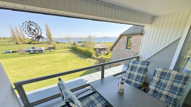 A balcony with a table and seating, overlooking a grassy area with a house and a body of water in the distance. - Apartment 06 with balcony and view of the Bodden - MR: Haus Inselperle with view of the (Mönchgut)