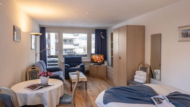 A hotel room with a bed, a desk with a chair, a sofa, a TV, a wardrobe, and a window with a view of buildings. - Chesa Corvatsch 111 - Champfer by Interhome (Champfer)