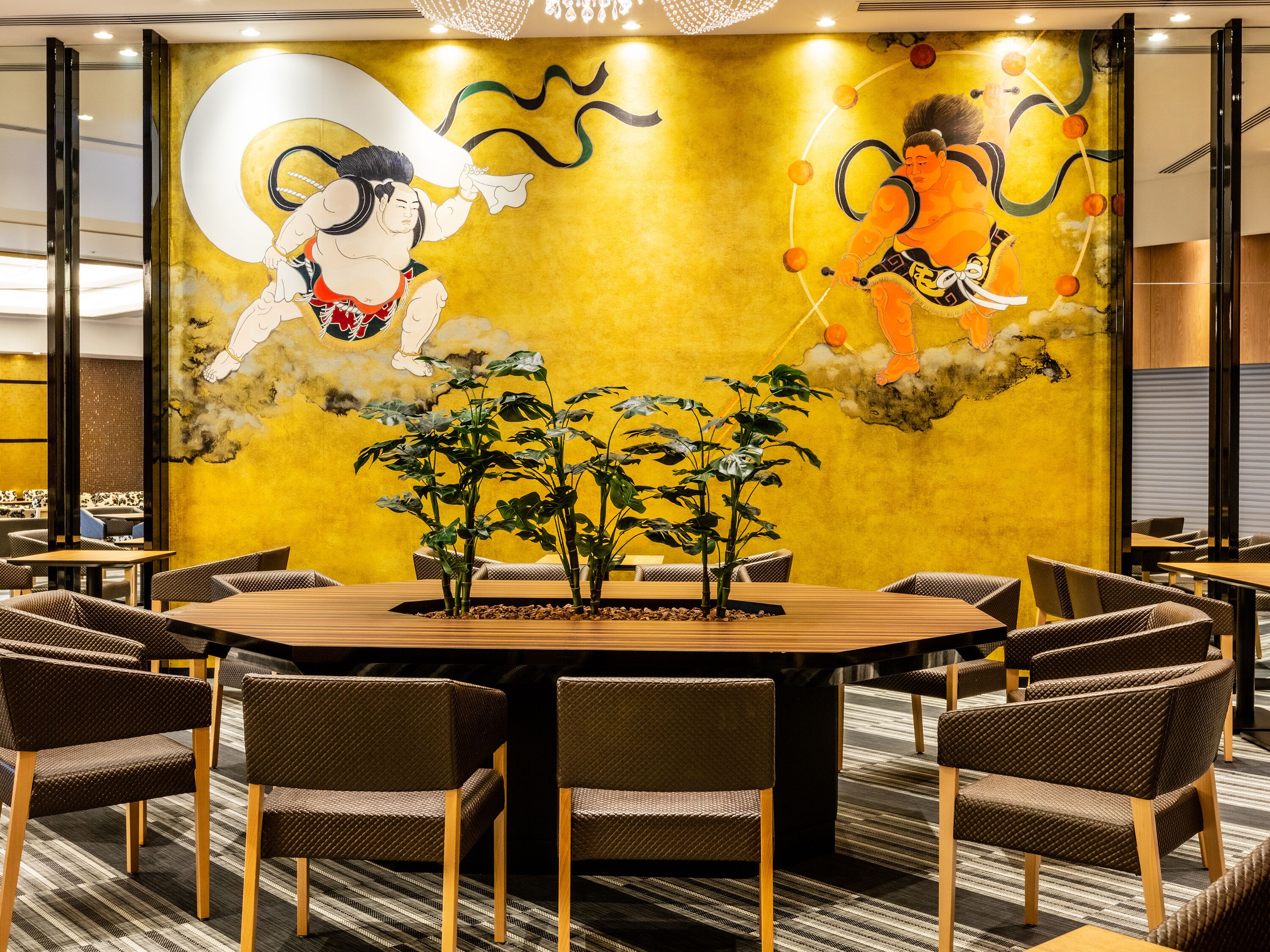A restaurant with a round table surrounded by wicker chairs, a yellow mural with traditional figures, and indoor plants.