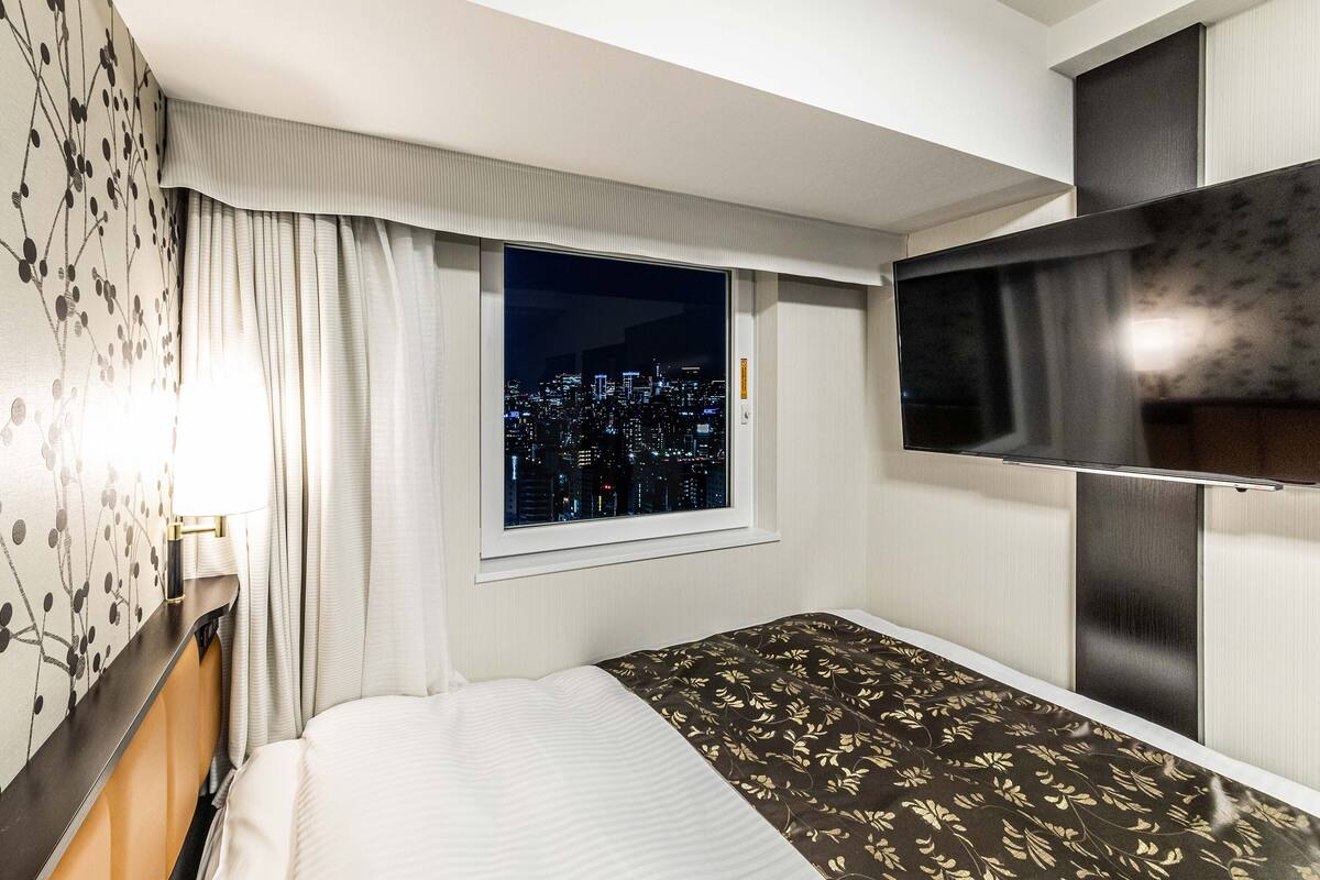 A hotel room with a bed, a TV, a window with a city view, and a curtain.