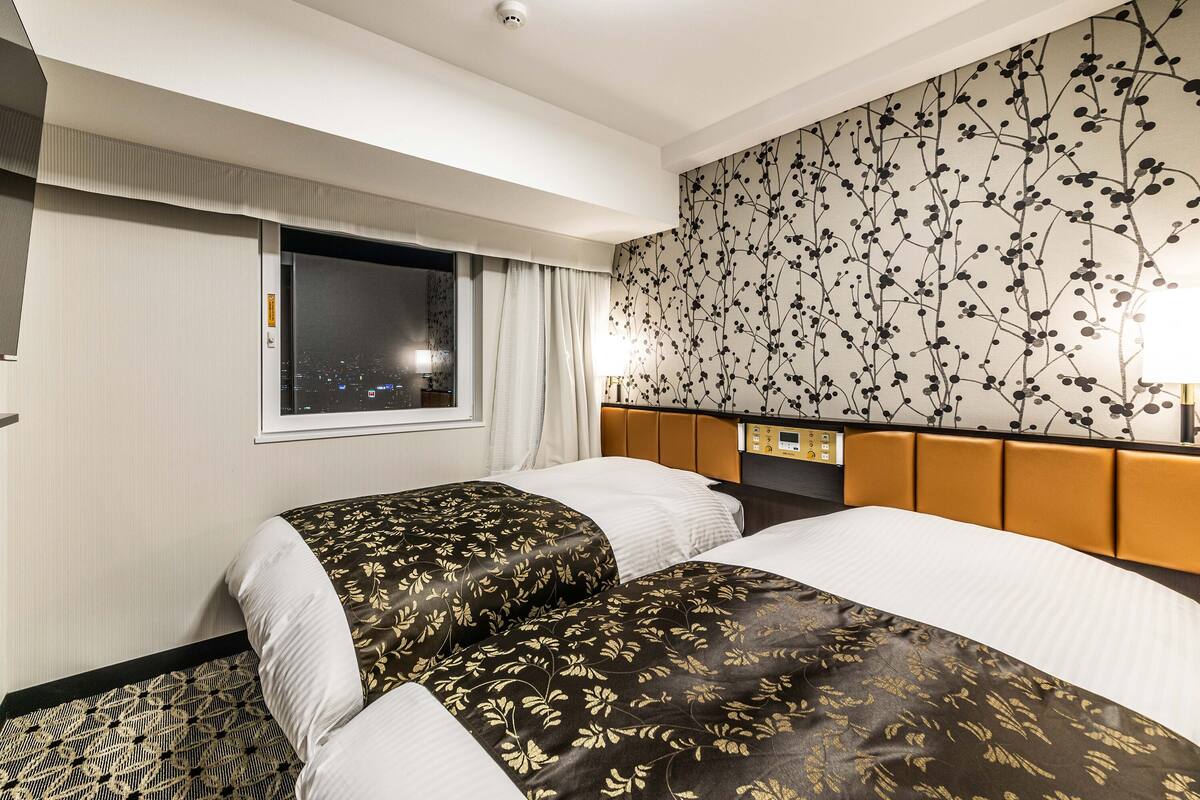 A hotel room with two beds, a window with a view of the city at night, a wall with a floral pattern, and a control panel on the headboard.