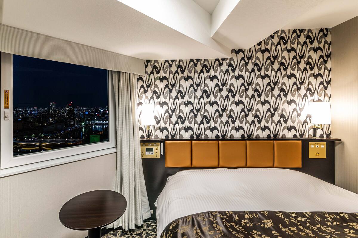 A hotel room with a city view, a bed, a bedside table, and a headboard with a patterned design.