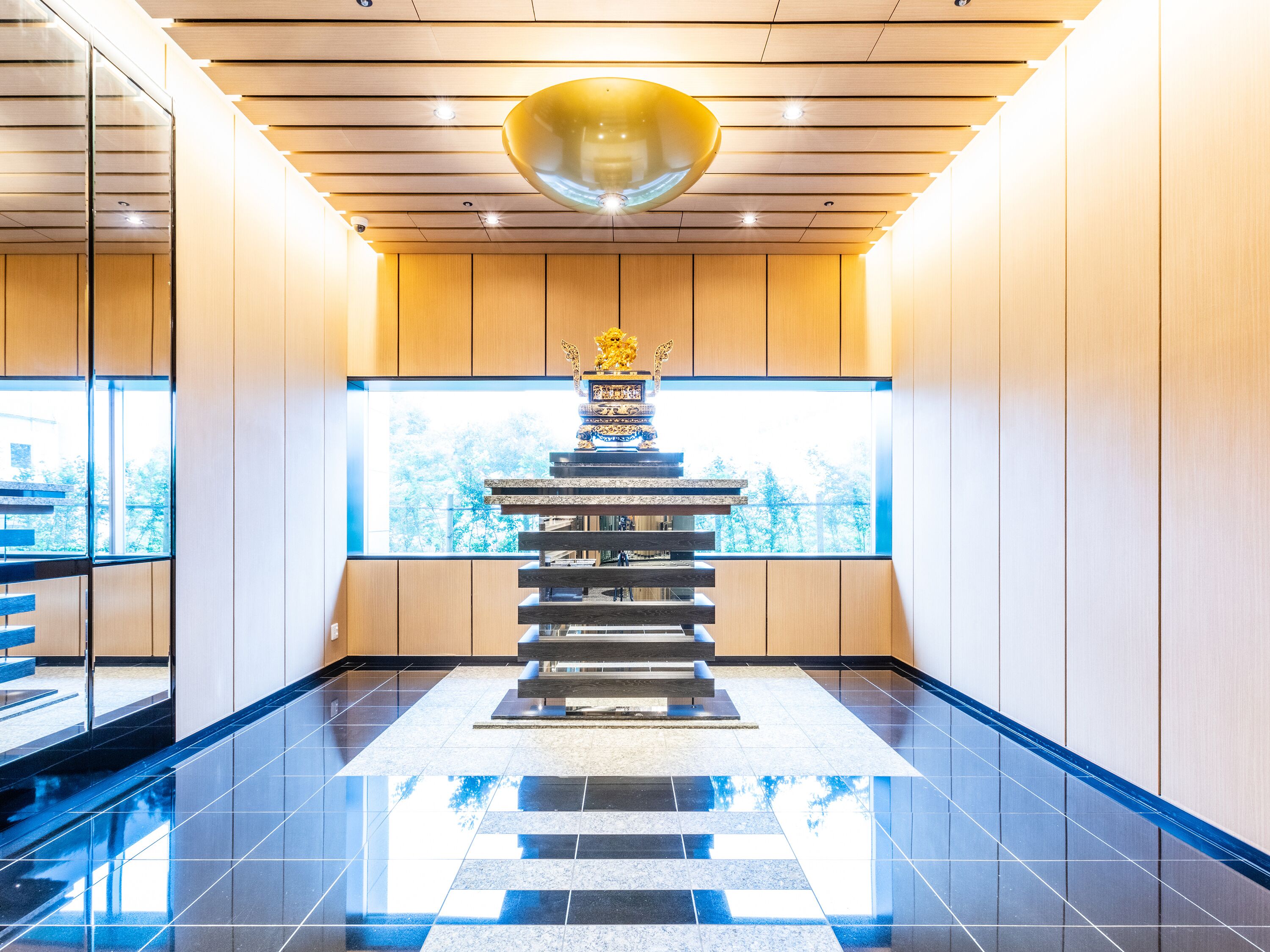 A modern interior with a reflective ceiling, a central staircase, and a large window offering a view of trees.