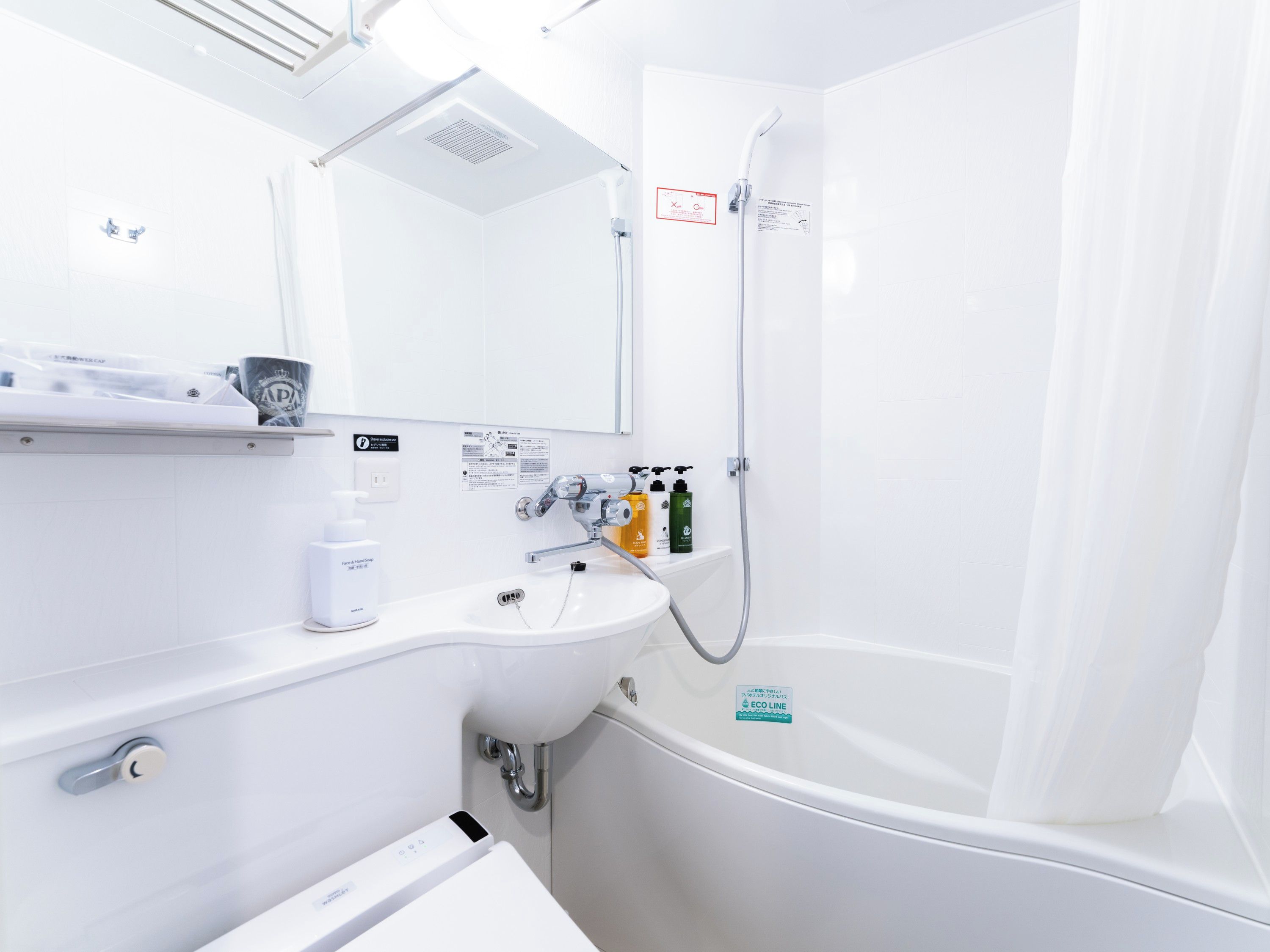 Une salle de bain moderne avec un lavabo blanc, un miroir, une douche et des toilettes. On trouve des articles de toilette sur le comptoir du lavabo.
