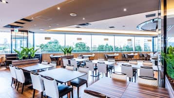 Un restaurant moderne avec des tables et des chaises en bois, de grandes fenêtres donnant sur la ville et des plantes d’intérieur.