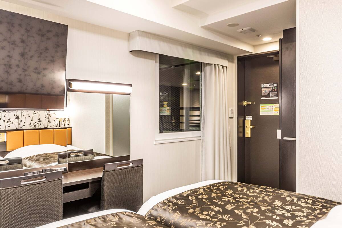A hotel room with a bed, a desk with a chair, a mirror, and a window with curtains.