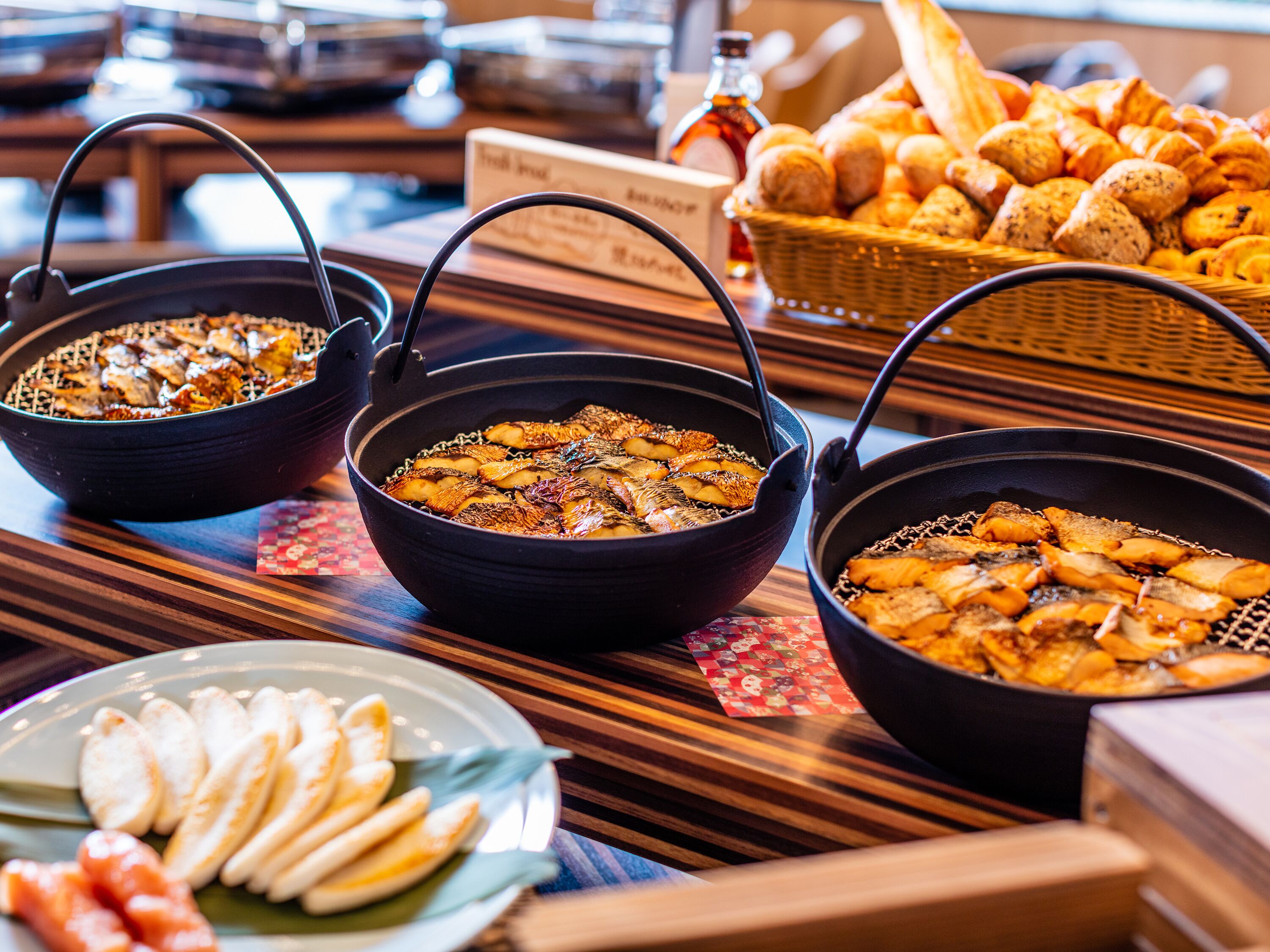 A buffet table with three black bowls containing food, a basket of bread, and a plate of dumplings.