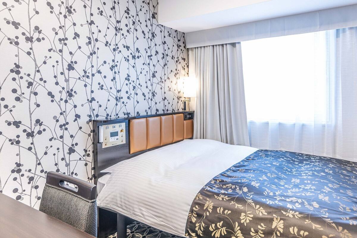 A hotel room with a bed, a control panel, a window with curtains, and a wall with a floral pattern.