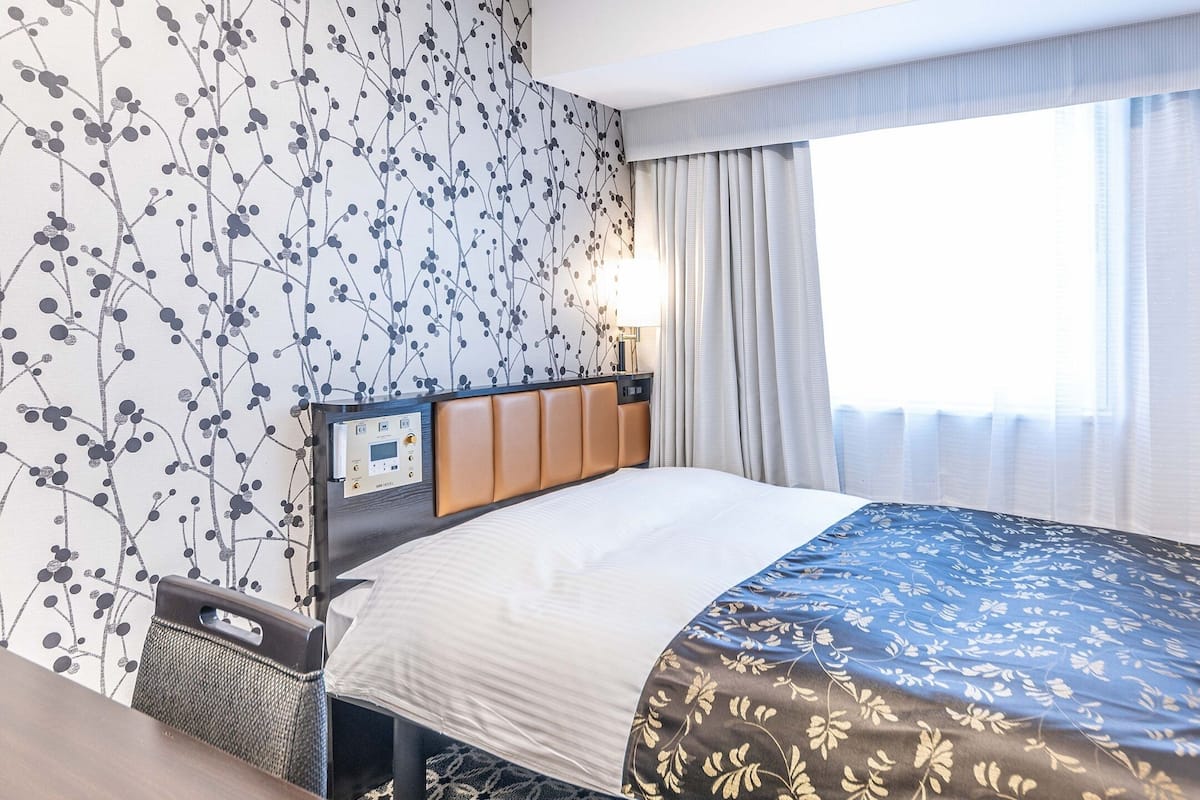 Una habitación de hotel con una cama, un panel de control, una ventana con cortinas y una pared con diseño floral.