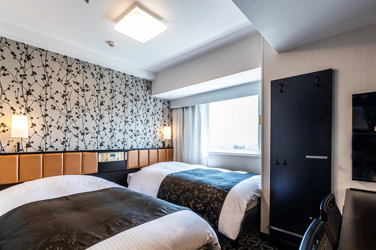 A hotel room with two beds, a headboard with a control panel, a window with a view, and a wardrobe.
