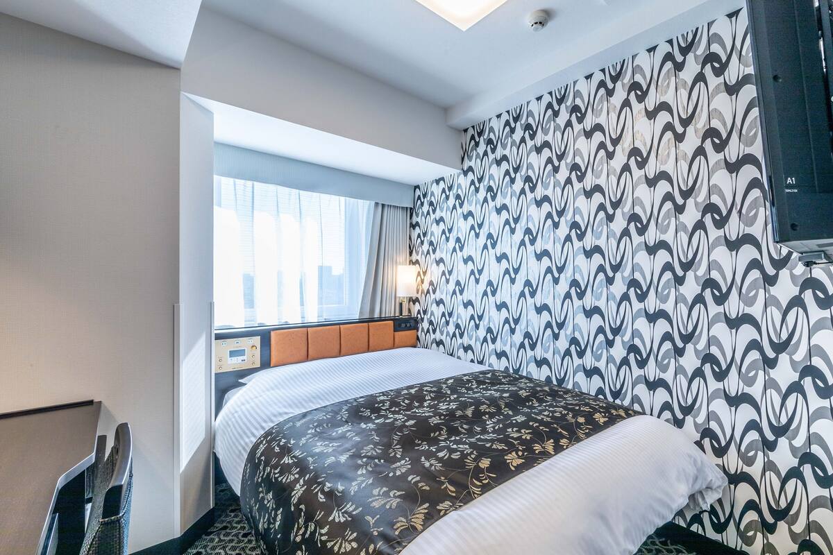 A hotel room with a bed, a window with curtains, a patterned wall, and a television.