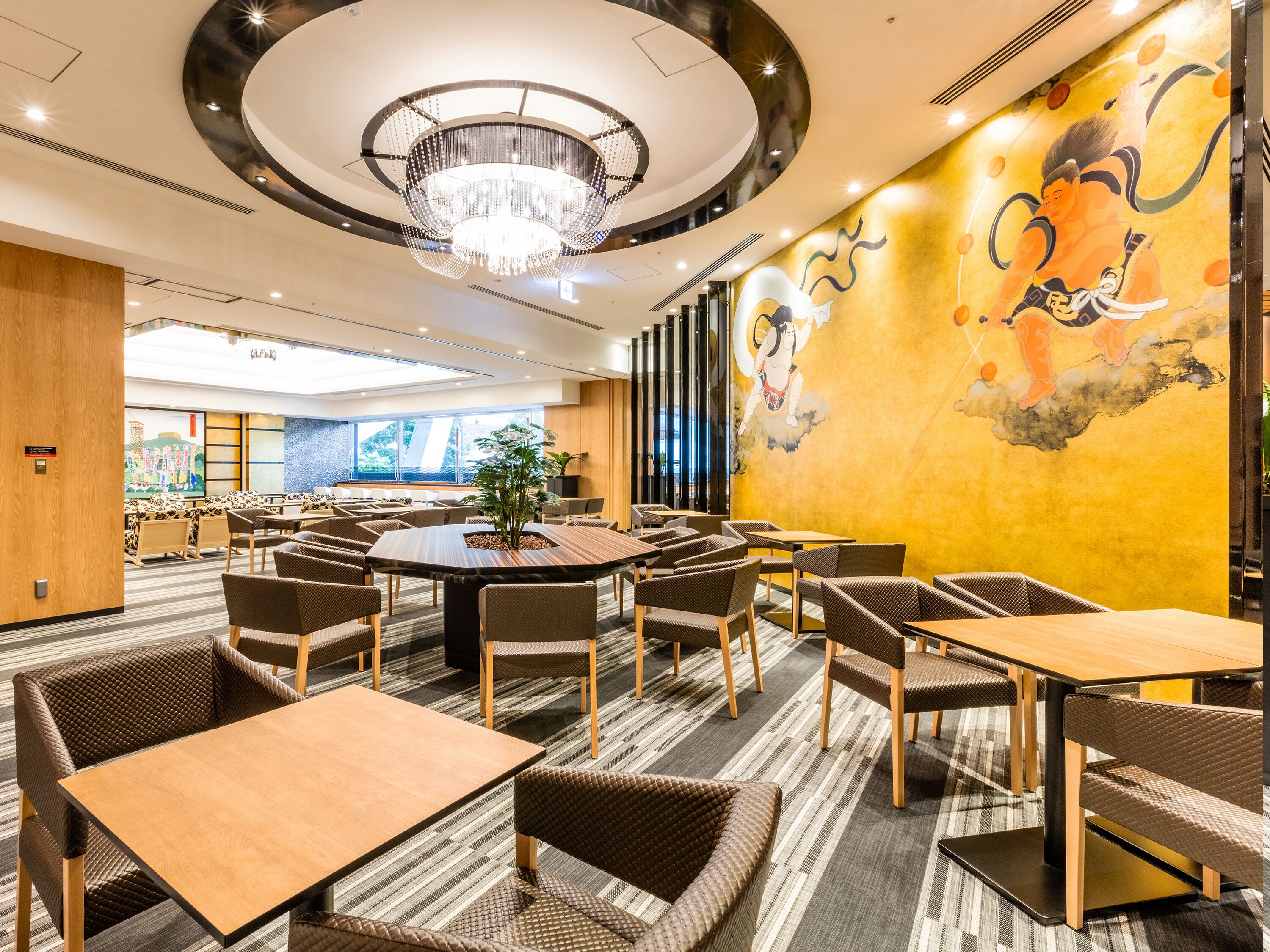 A modern hotel lobby with a large round table, multiple seating areas, and a mural on the wall.