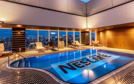 A modern indoor swimming pool with a city view, surrounded by lounge chairs and a checkered floor pattern.