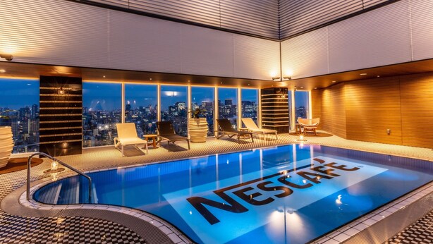 Piscina interior moderna con vistas a la ciudad, rodeada de sillas de descanso y un patrón de suelo a cuadros.