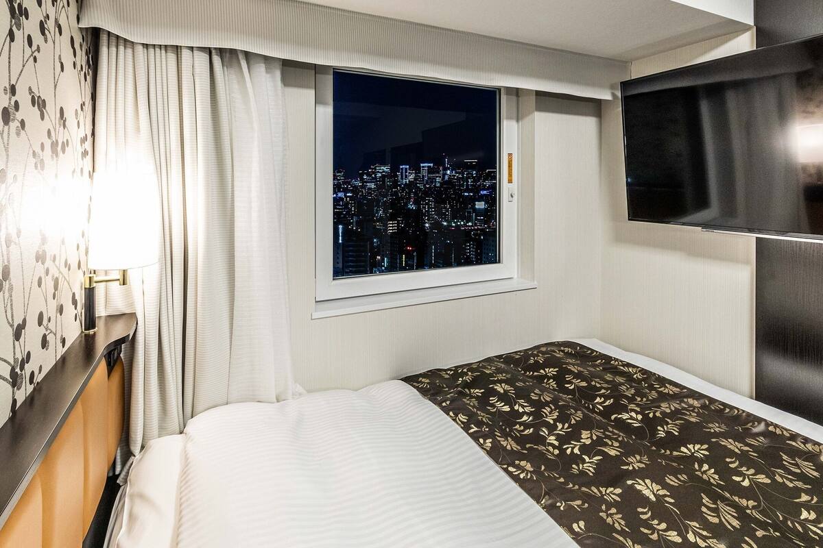 A hotel room with a bed, a window with a city view, a TV, and a lamp.