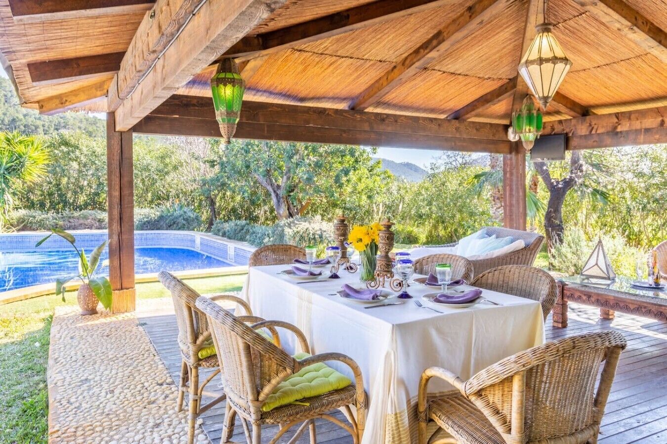 Un espace extérieur aménagé pour les repas, avec une pergola couverte, des chaises en osier, une table dressée pour deux personnes et une piscine en arrière-plan.