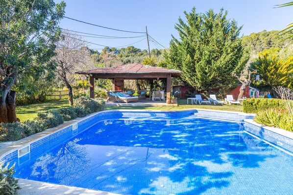 Une piscine dotée d’une terrasse carrelée, d’un espace salon couvert et entourée de verdure.