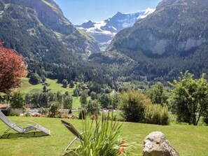 A mountainous landscape with a valley, a lawn with a lounge chair, and a rock.
