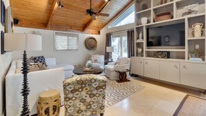A modern living room with a ceiling fan, white sofas, a patterned armchair, a coffee table, and a bookshelf with decorative items.