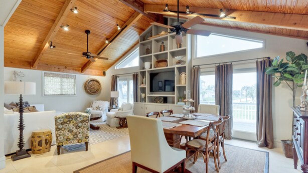 A spacious living area with a high ceiling, wooden beams, a dining table with chairs, a television, and a large window with curtains.