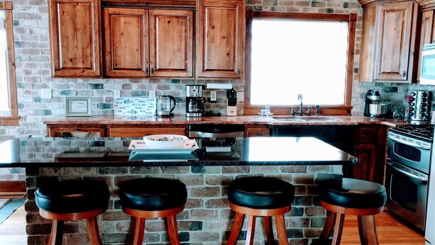 A kitchen with wooden cabinets, a brick wall, and a large window. There are three stools and a sink. - Amazing view - Lake House Retreat with Mountain Cabin Warmth (Garden City)