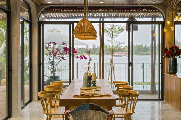 Un espace repas avec une table et des chaises en bois, un luminaire suspendu et une vue sur une étendue d’eau grâce à de grandes fenêtres.