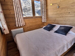 Quarto de madeira com cama, cortinas, radiador e janela com vista para uma paisagem nevada.