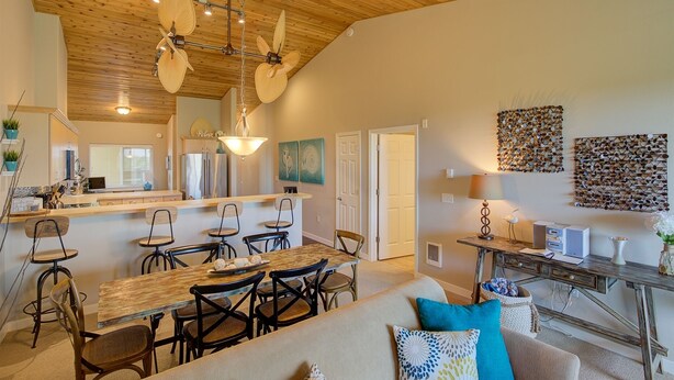 A spacious living area with a wooden ceiling, a dining table with chairs, a sofa with cushions, and a kitchen area with a refrigerator and a bar counter.