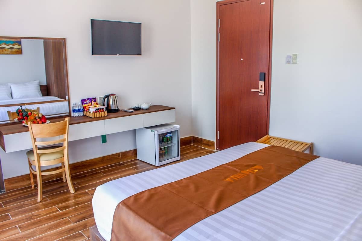 A hotel room with a bed, a desk with a chair, a television, a wardrobe, and a small refrigerator.