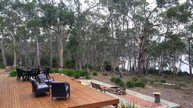 Eine Holzterrasse mit Gartenmöbeln, umgeben von einer natürlichen Landschaft mit Bäumen und einem Gewässer im Hintergrund.