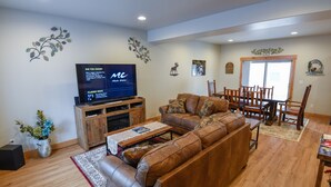 A living room with a brown leather sofa, wooden dining table, and chairs. A flat-screen TV is mounted on the wall.