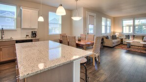 A spacious kitchen with a large island, wooden flooring, and a dining area with wicker chairs. - Beach Tripping - Oyhut Bay Seaside Village (Ocean Shores)