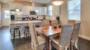 A modern kitchen with white cabinets, stainless steel appliances, a wooden dining table, wicker chairs, and pendant lighting. - Beach Tripping - Oyhut Bay Seaside Village (Ocean Shores)