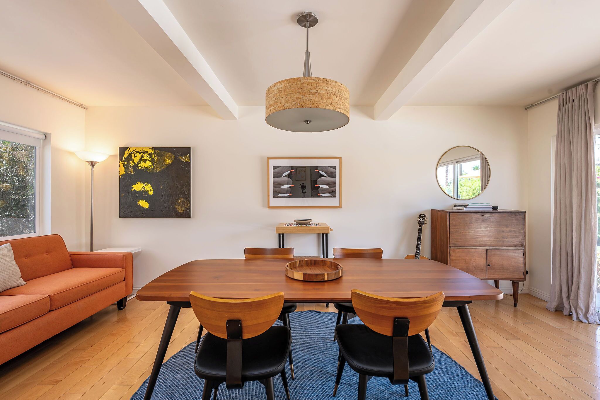 A dining area with a wooden table, chairs, a hanging light fixture, a sofa, a cabinet, and artwork on the walls.