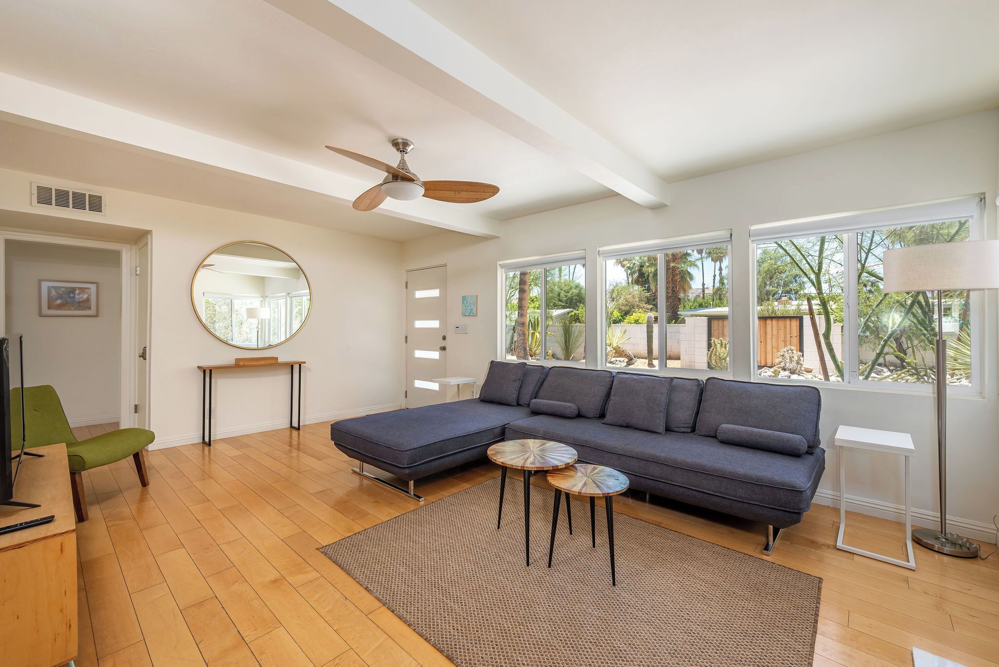 Un salon moderne avec un canapé d’angle, un grand miroir rond, un ventilateur de plafond et de grandes fenêtres.